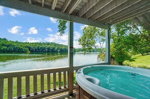 A hot tub on a covered deck with a view of a lake and forest. - Lakefront Home: Fishing + Kayaking + King Bed + Hot Tub (Logan)