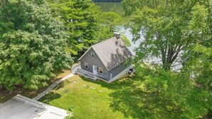 A two-story house with a sloped roof, surrounded by lush greenery and a lake. - Lakefront Home: Fishing + Kayaking + King Bed + Hot Tub (Logan)