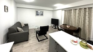 A modern living room with a sofa, a TV, a dining table, and a kitchen area. - Cozy 2-Bedroom Condo with Parking in Niagara Falls (Niagara Falls)
