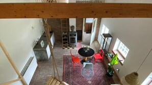 A high-ceilinged room with exposed wooden beams, a central seating area with a yellow sofa, red and blue chairs, and a hanging chair. A brick fireplace is visible in the background.