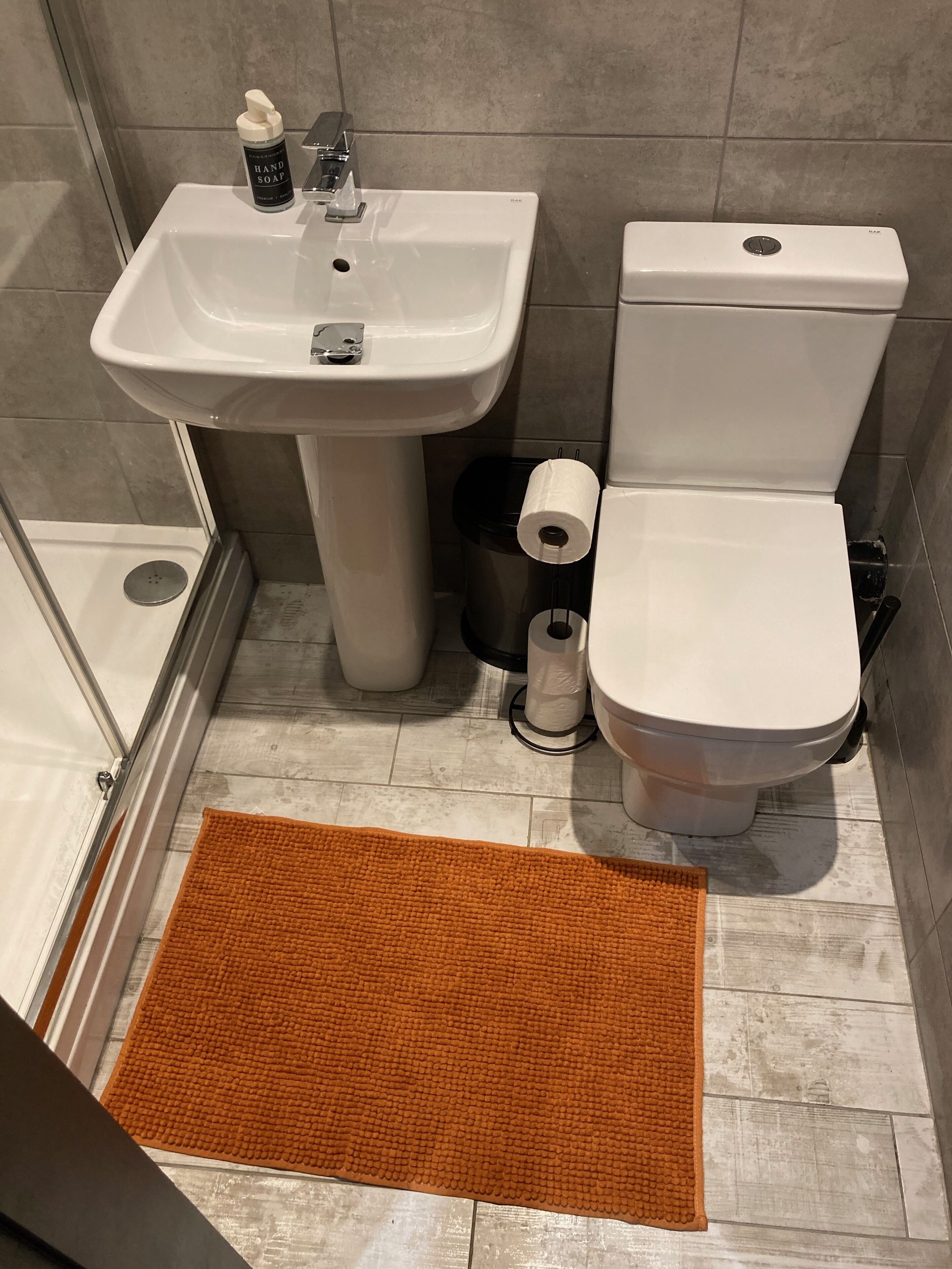 Une salle de bain avec un lavabo, des toilettes et une douche, tous de couleur blanche. On peut voir un rouleau de papier toilette.