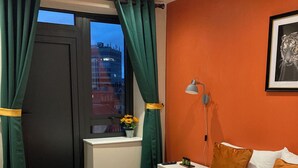 Una camera d'albergo con un letto, tende verdi, una finestra che si affaccia sulla città, un comodino, una lampada e un quadro appeso al muro.
