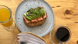 En morgenmadsopsætning med en tallerken med laks og avocado på ristet brød, en skål med frugt og to kopper kaffe på et træbord.