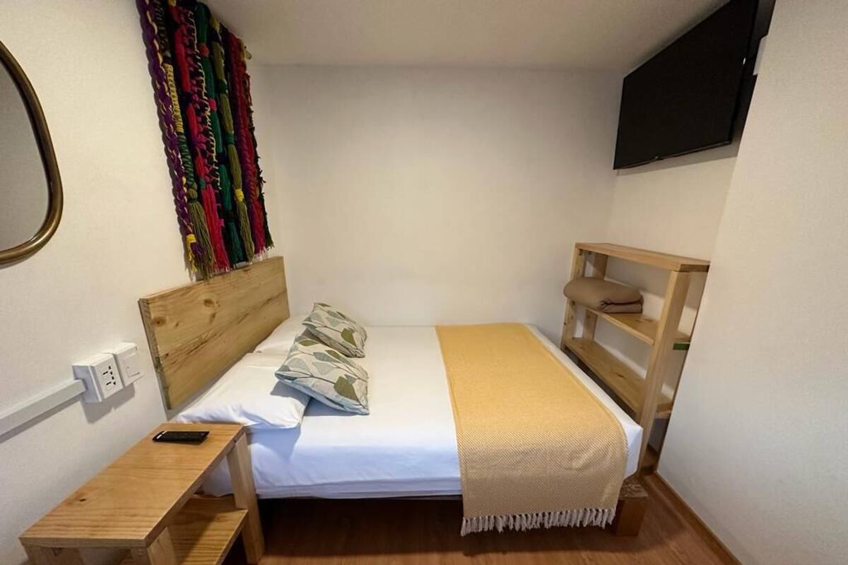 Une petite chambre à coucher, bien rangée, avec un lit simple, une table de chevet, un téléviseur fixé au mur et une tapisserie aux couleurs vives.