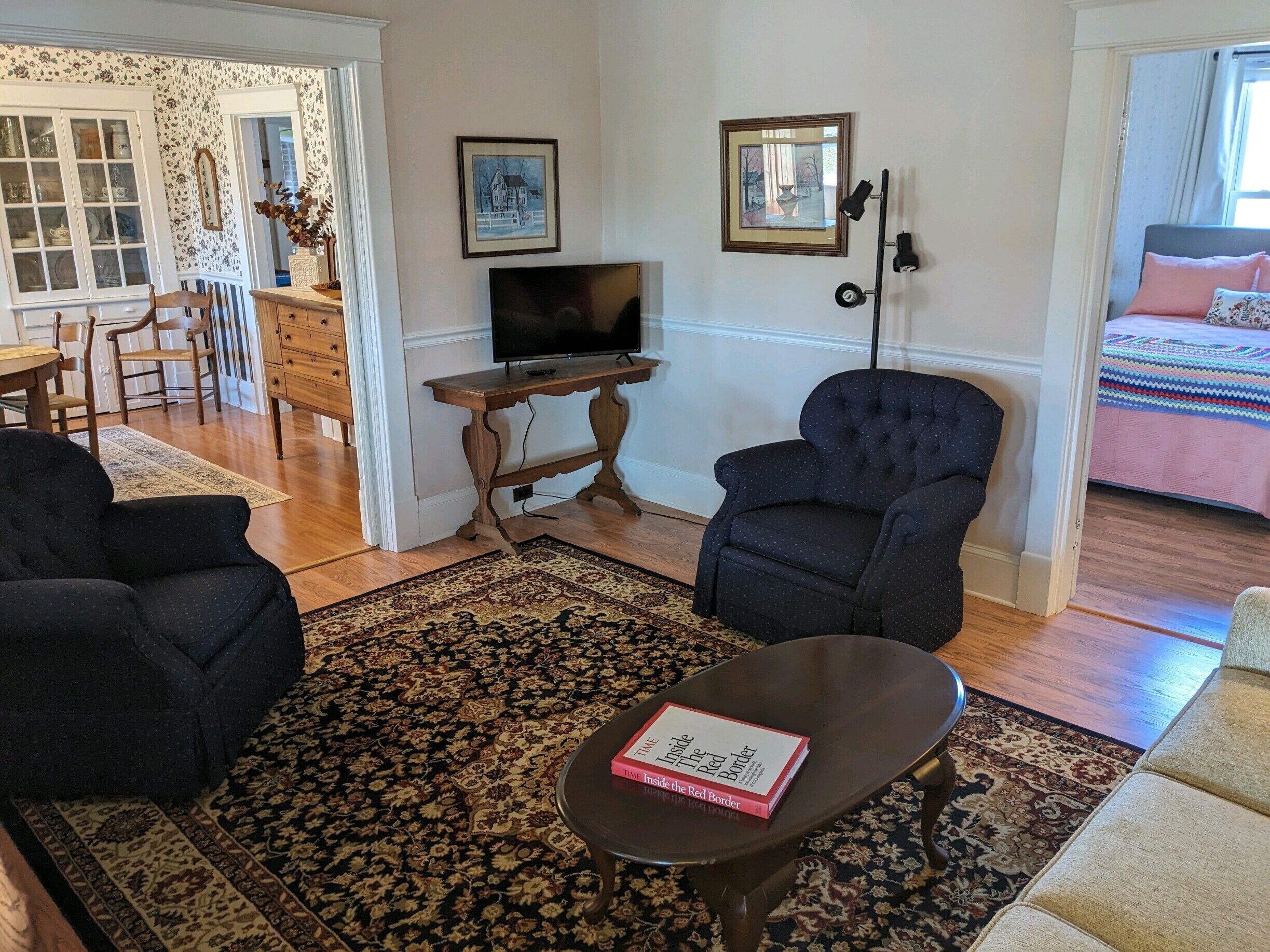 A living room with a television, two armchairs, a coffee table with a book, a patterned rug, and a bed with a colorful blanket in the background.