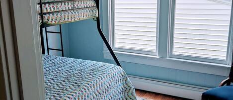 Un dormitorio con una cama alta, una ventana con persianas, una alfombra estampada y una silla.