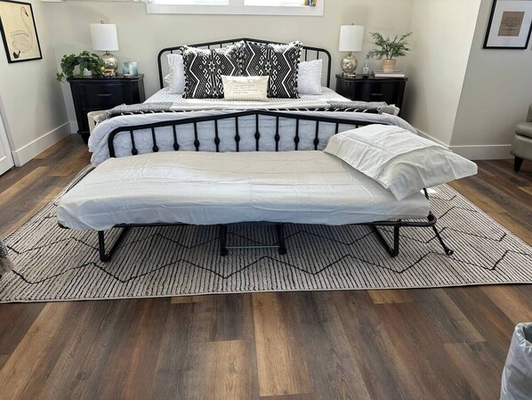 A convertible bed in a bedroom with a geometric-patterned rug, wooden flooring, and bedside tables with lamps and plants.