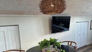 Un coin repas avec une table ronde noire, deux chaises en bois, un luminaire suspendu en fibres tressées, une plante en pot et un téléviseur fixé au mur.