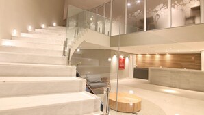 A modern interior with a glass staircase, a round wooden table, and a seating area with a cushioned chair. - hihome - Royale (Cabo Frio)