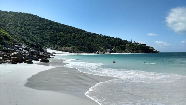 A beach with clear turquoise water, a sandy shore, and a forested hill in the background. - hihome - Royale (Cabo Frio)