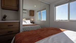 Une chambre avec une grande fenêtre, un lit recouvert d’une couverture marron, une table de chevet avec une lampe et une armoire en bois.