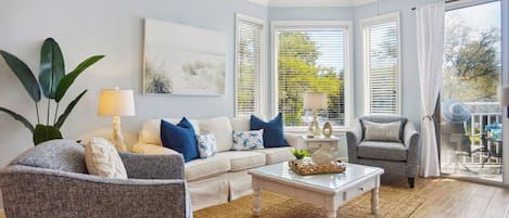 Una sala de estar con sofá, sillón y una mesa de centro, un ventilador de techo y vistas a través de amplios ventanales.