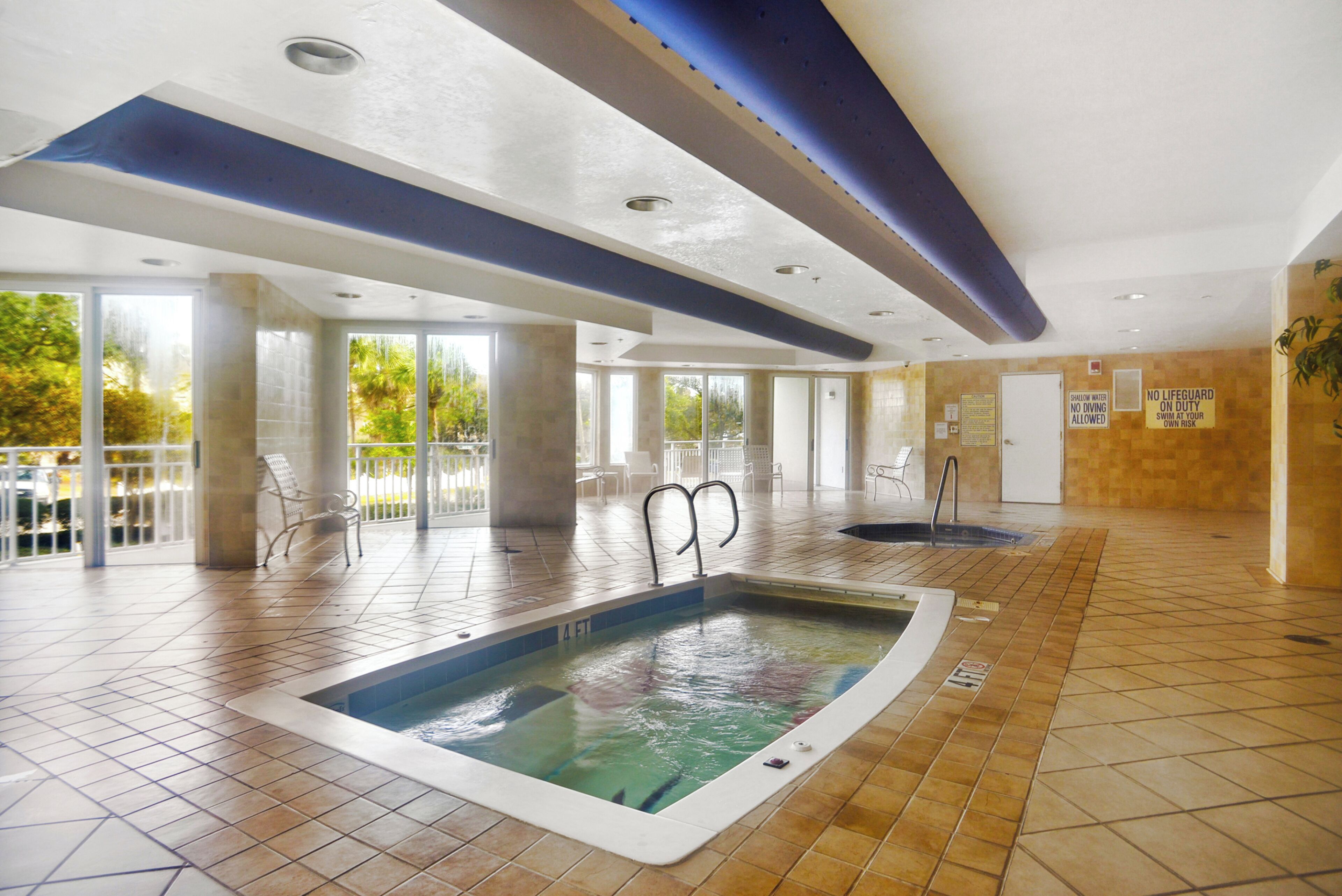 An indoor swimming pool with a view of the outdoors, surrounded by chairs and a tiled floor.