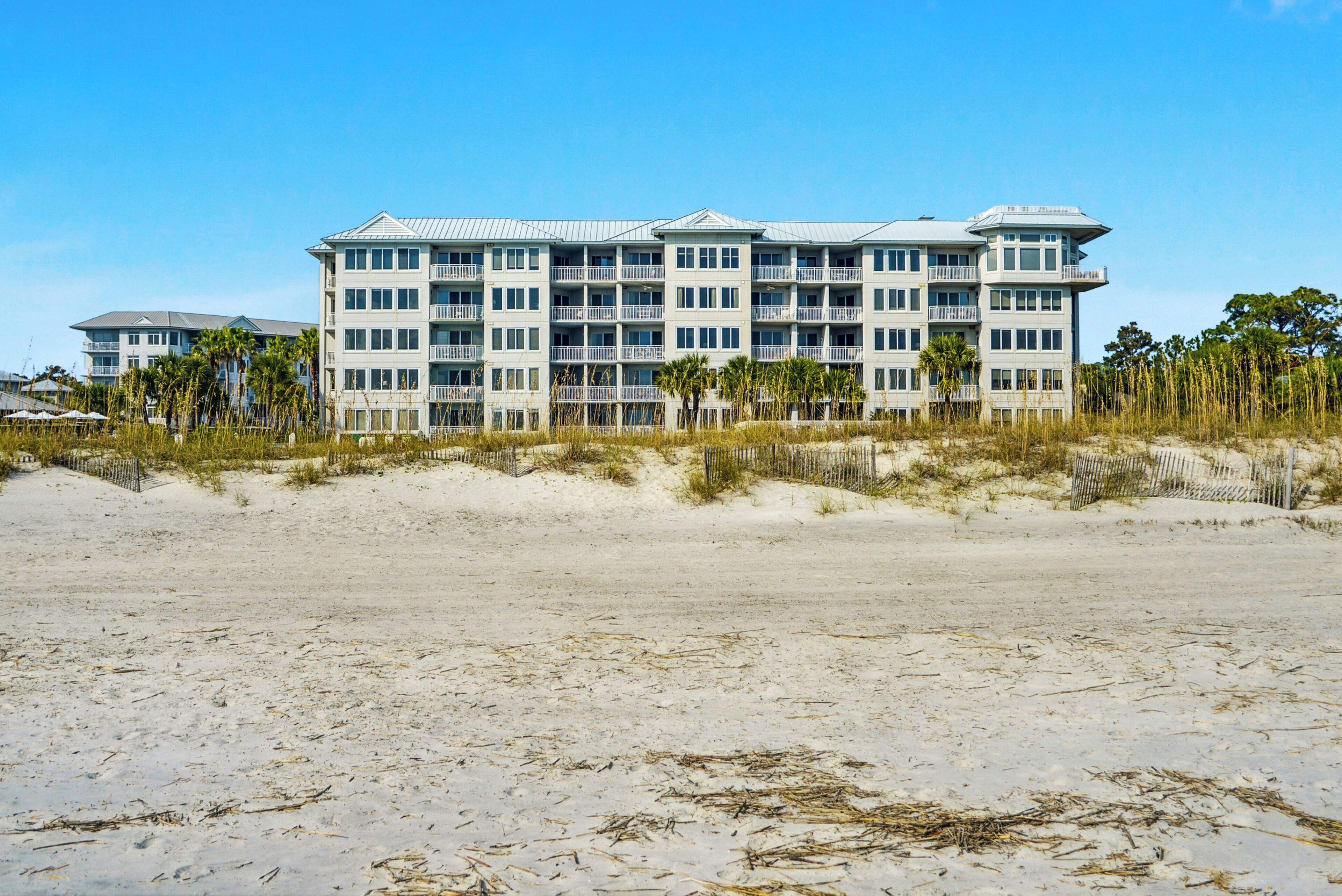 A beachfront hotel with multiple balconies, a sandy beach in the foreground, and palm trees.