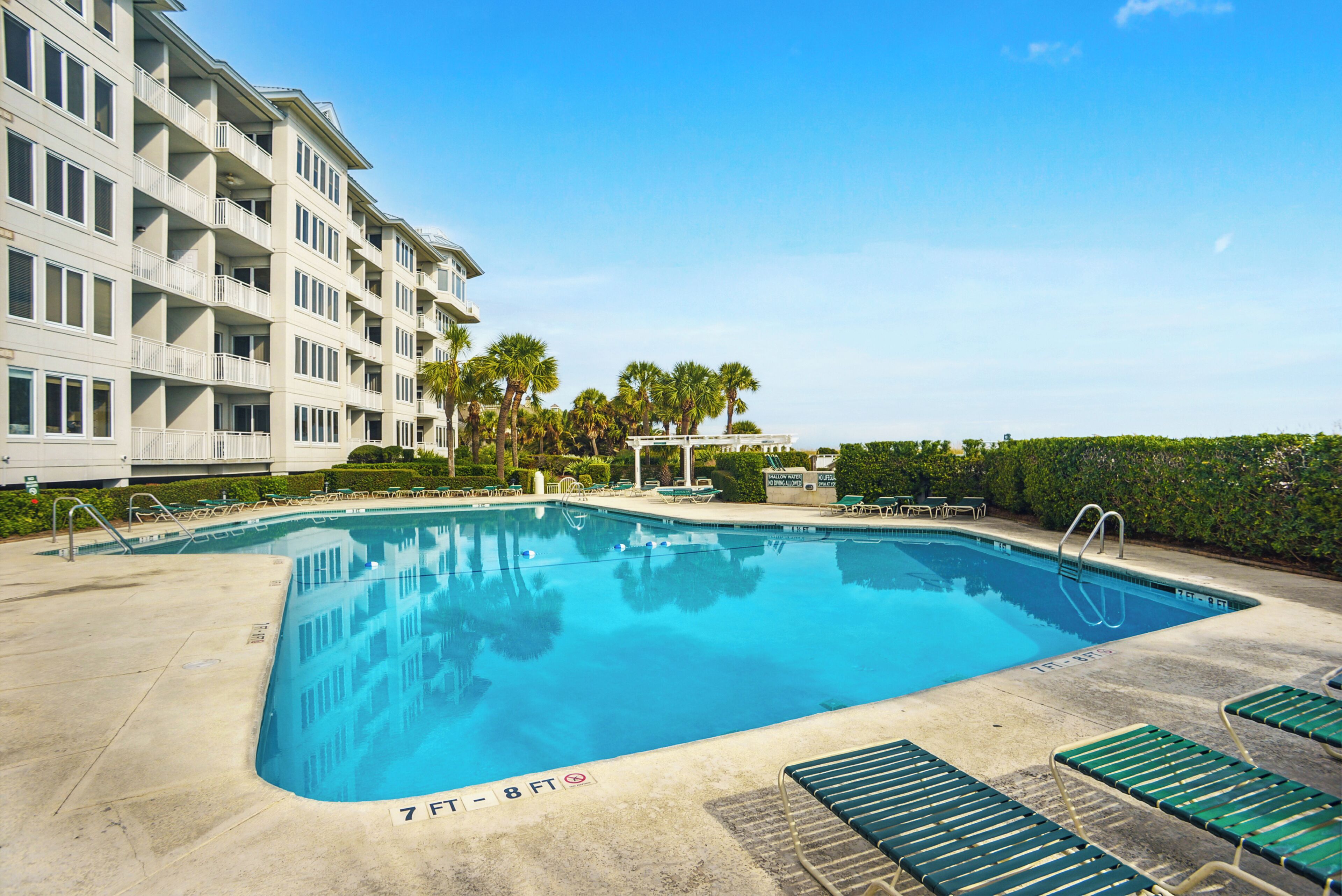 A rectangular swimming pool with a clear blue water surface, surrounded by a concrete deck. There are lounge chairs and a small bridge over the pool.