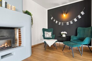 A modern living room with a fireplace, two green armchairs, a small table with a vase of flowers, and a wall-mounted light fixture.