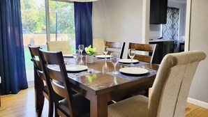 A dining area with a wooden table set for four, surrounded by chairs. There is a chandelier above the table, and a view of the outdoors through large windows. - 5BD Cozy, SouthGlenn Gem, Yard, Games, Shops, close to Downtown Littleton&Denver (Centennial)