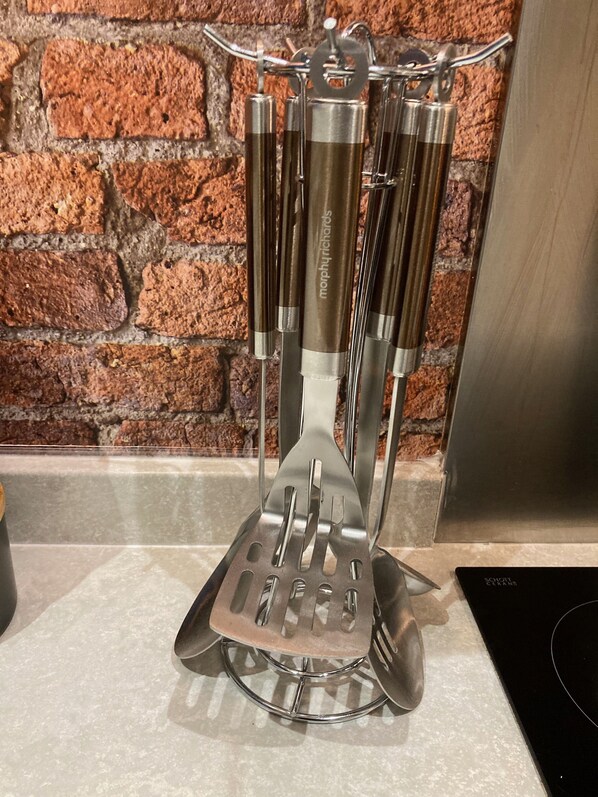 Un recipiente para utensilios de cocina con varias espátulas y un rallador, sobre una encimera frente a una pared de ladrillo.