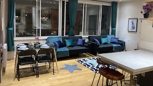 Ein modernes Wohnzimmer mit einer Couch, einem Esstisch, Stühlen und großen Fenstern mit Vorhängen.