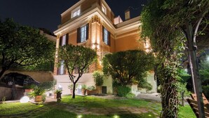 A well-lit courtyard with a stone pathway, surrounded by lush greenery and a large building with multiple windows. - Villa Michelangelo, your home in the heart of the eternal city (Roma)