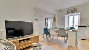Ein modernes Wohnzimmer mit Flachbildfernseher, eine Essecke mit Stühlen und Tisch sowie eine Küche mit Geschirrspüler.