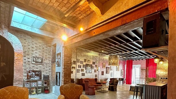 A spacious lounge area with a high ceiling, exposed brick walls, and a variety of seating options including armchairs and a round table.
