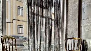 A room with two wooden chairs and a small round table, a curtain with vertical stripes, and a view of buildings outside.