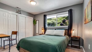 Ein Schlafzimmer mit einem Bett, einem Schreibtisch und einem Stuhl. Ein Fenster mit Vorhängen, eine Pflanze und ein Bild an der Wand sind ebenfalls vorhanden.