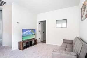 Un salón con un televisor de pantalla plana sobre un soporte de madera, un sofá gris y una puerta que da a otra habitación.