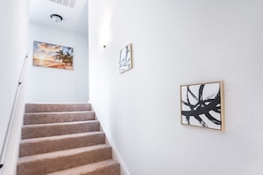 Una escalera con peldaños cubiertos de moqueta color beige, paredes blancas y tres obras de arte enmarcadas.