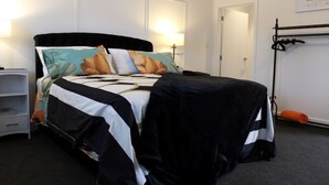 A neatly made bed with a black headboard, a bedside table with a lamp, and a coat rack with a folded jacket. - The Pool House Bed & Breakfast - 50 sq Kitchenette Studio - Near Marine Parade (Napier)