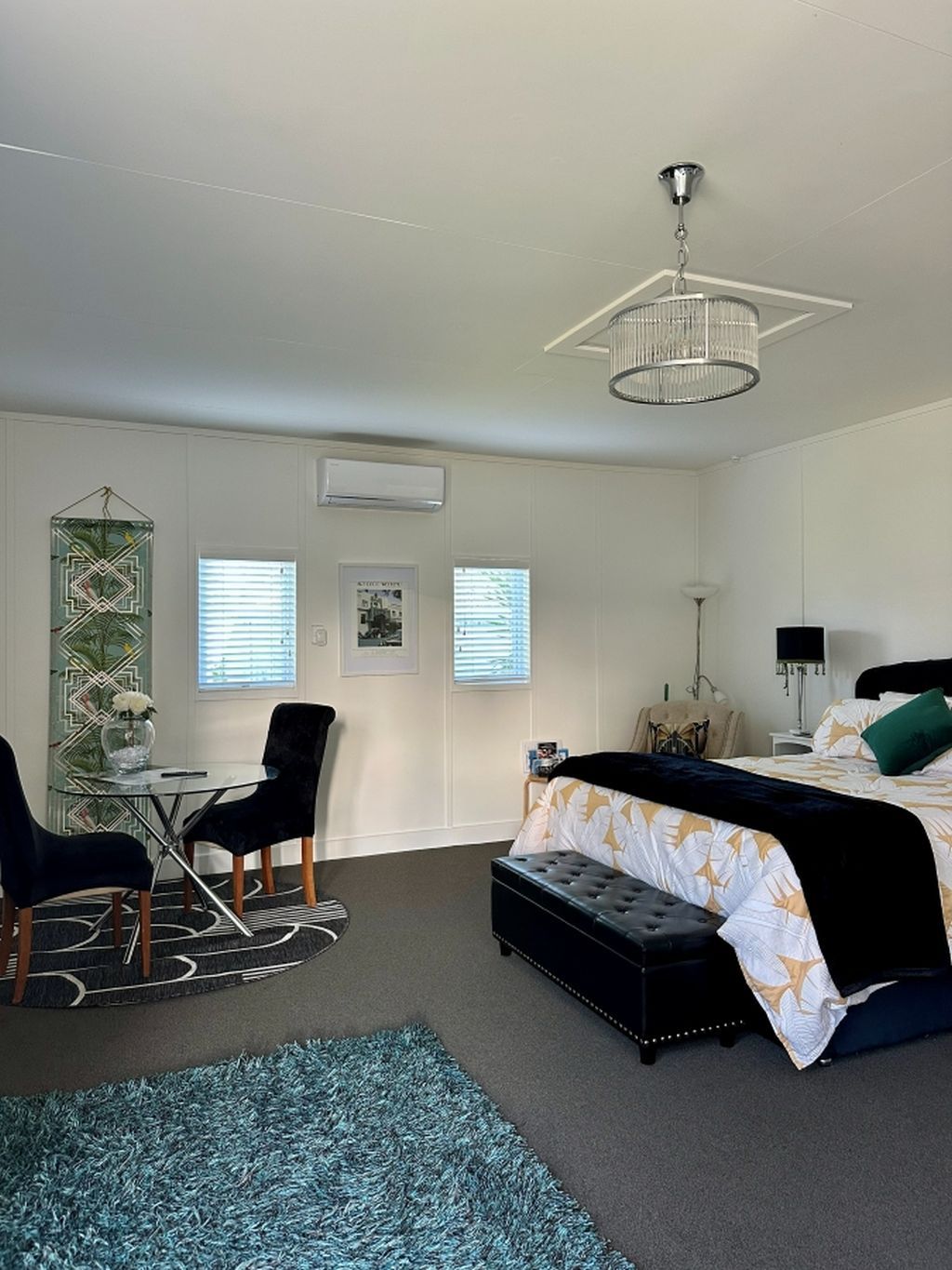 A hotel room with a bed, a desk with a chair, a small table, and a large window with blinds.
