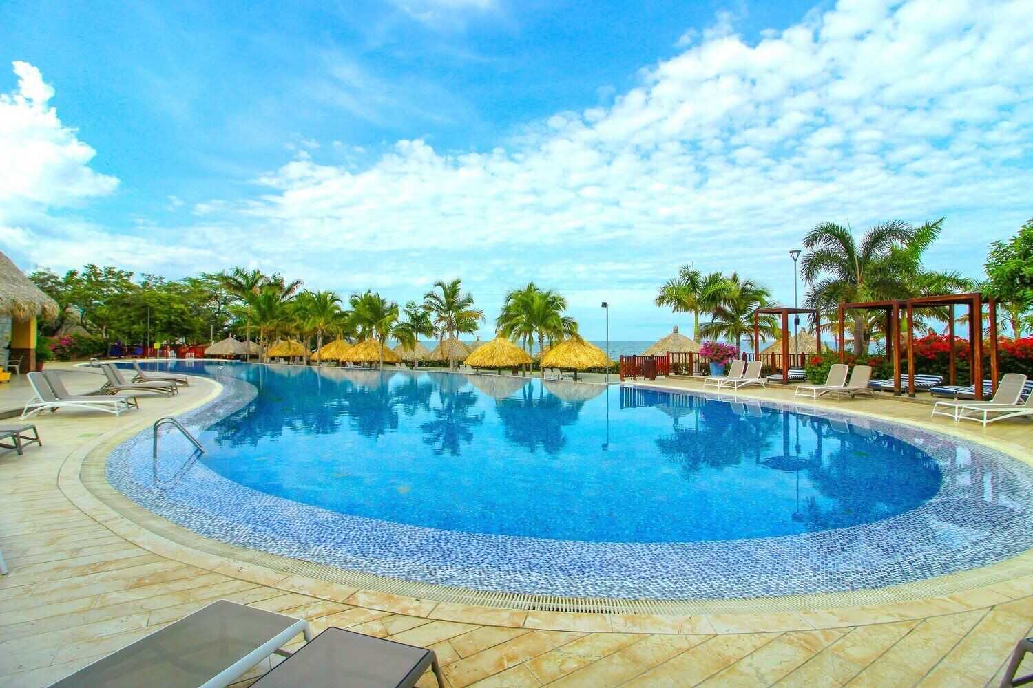 Ein kreisrunder Pool mit Liegestühlen und Strohdächern, umgeben von Palmen und einem klaren Himmel.