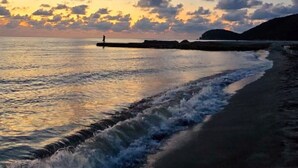 A beach scene with a person standing on a pier, waves crashing onto the shore, and a sunset in the background. - Luxurious Sea View Apartment - Samaria Club (Gaira)