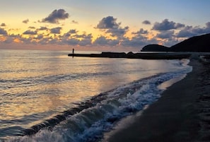 A beach scene with a person standing on a pier, waves crashing onto the shore, and a sunset in the background. - Luxurious Sea View Apartment - Samaria Club (Gaira)