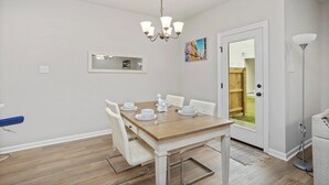 Uma área de refeições com mesa de madeira, cadeiras brancas, um lustre e uma porta que dá para o exterior.