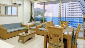 A modern living room with a sofa, chairs, a glass coffee table, and a dining table. There is a large window with a view of buildings and mountains. - Amazing Apartment in Beach Club - Samaria! (Gaira)