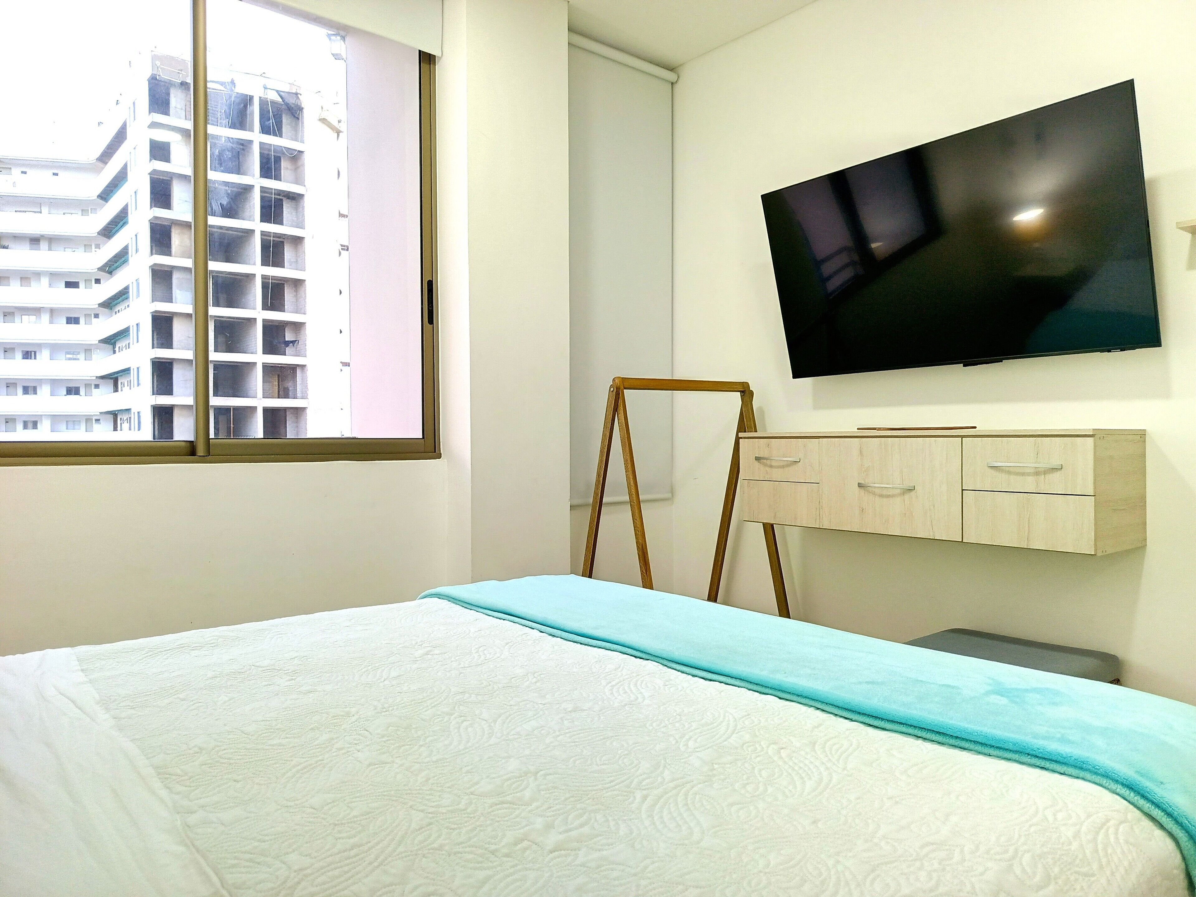 A hotel room with a bed, a TV mounted on the wall, a window with a view of a building, and a wooden dresser with drawers.