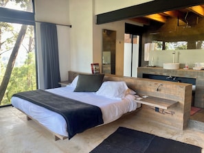 Un dormitorio moderno con una cama grande, una mesita de noche de madera, un banco empotrado, un espejo y vistas a árboles.