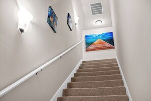 A carpeted staircase with a handrail, a painting of a wooden pier, and wall-mounted light fixtures. - Serene Oasis for Deep Relaxation Near LSU - CL5628 (Baton Rouge)