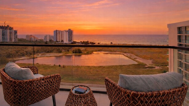 Un balcon avec des fauteuils en osier et une petite table, d’où l’on peut admirer un plan d’eau et des bâtiments au coucher du soleil.