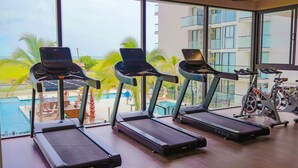 Une salle de sport équipée de tapis de course et de vélos d’appartement, avec vue sur une piscine et des palmiers.