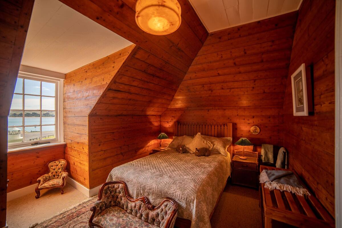 A cozy attic bedroom with a wooden sloped ceiling, a large bed, a chair, a nightstand, and a window with a view of the water.