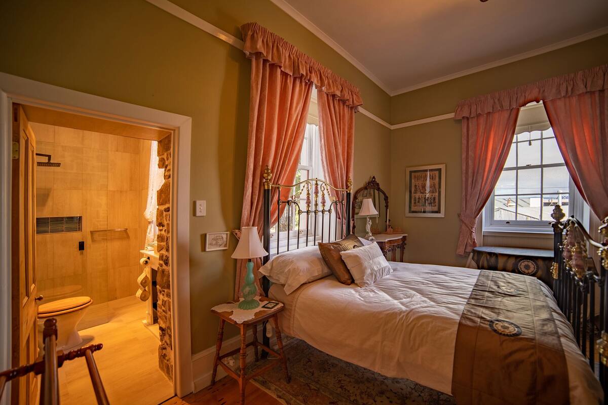 A bedroom with a large bed, a bedside table with a lamp, a window with curtains, and a bathroom with a shower and toilet.