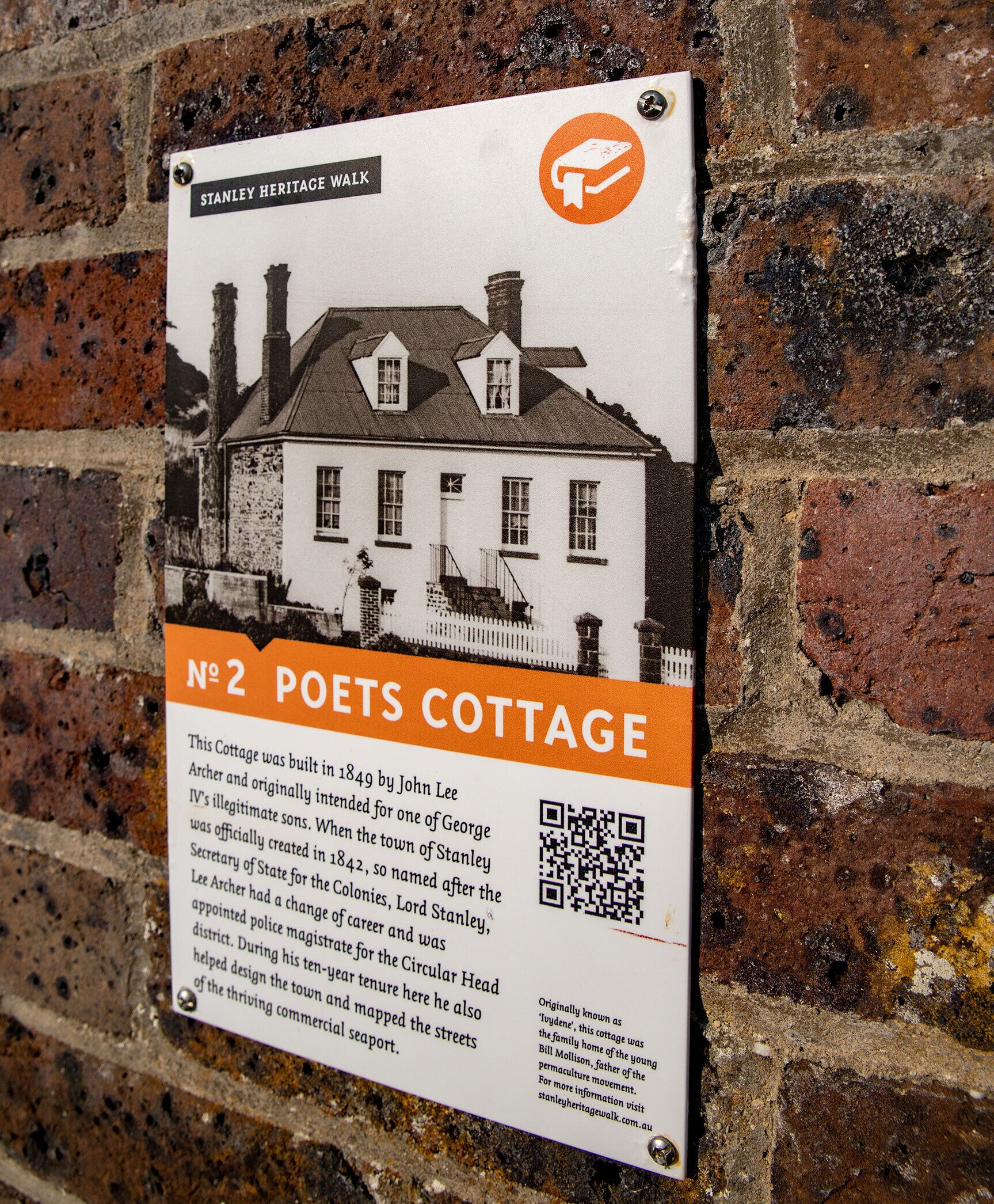A historical plaque on a brick wall, detailing the Poets Cottage, a building with a white facade and multiple chimneys, indicating its historical significance and architectural style.