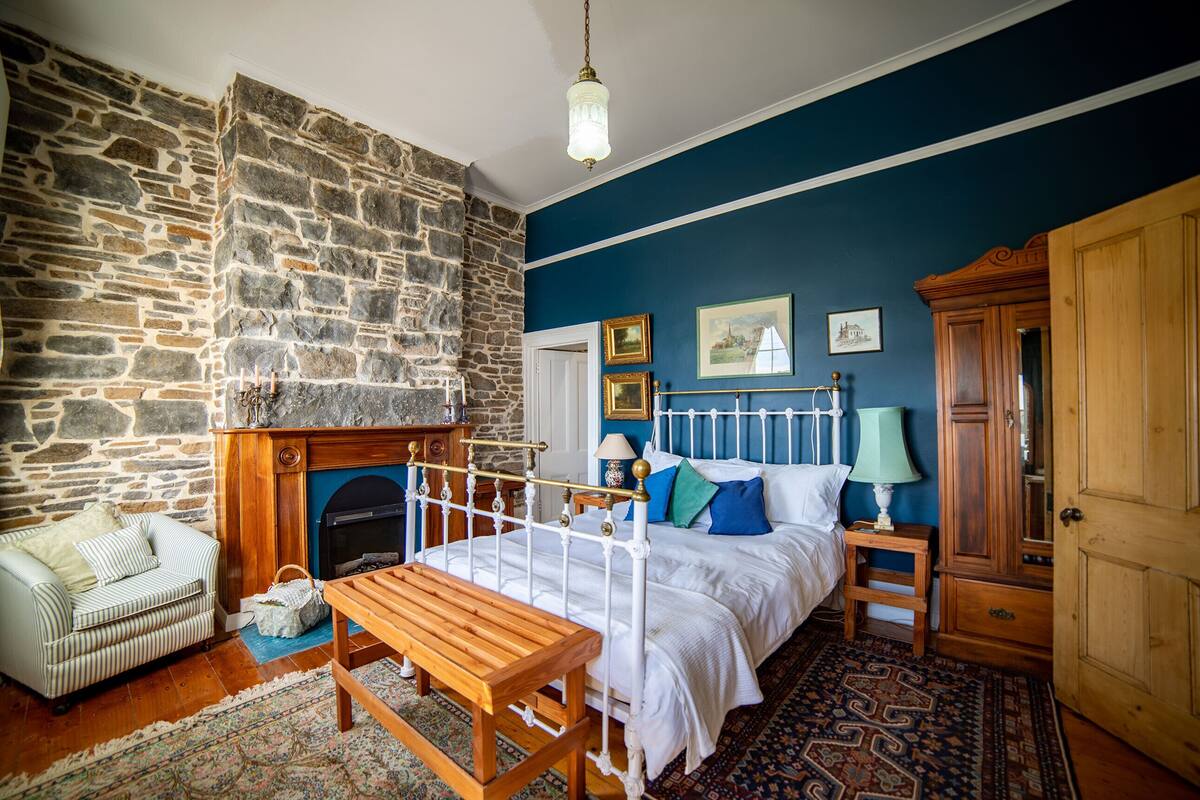 A bedroom with a stone fireplace, a bed with white bedding, a wooden bench, a patterned rug, and a wooden wardrobe.