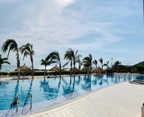 Une piscine bordée de palmiers et ombragée par des parasols en chaume, entourée de chaises longues et d’une terrasse en bois.