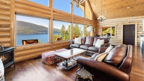 A log cabin interior with a large window overlooking a lake and mountains, furnished with leather sofas and a glass coffee table. - Ski, Swim, Relax at Whitefish Lake | Stunning Views of Whitefish Lake (Whitefish)