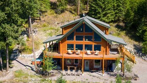 A log cabin with a large deck, multiple windows, and a stone foundation. - Ski, Swim, Relax at Whitefish Lake | Stunning Views of Whitefish Lake (Whitefish)