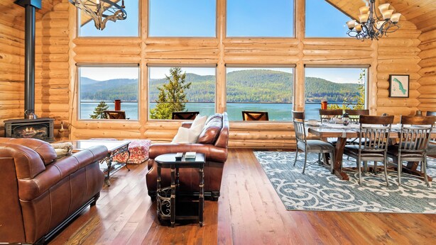 A log cabin interior with a fireplace, leather sofa, dining table, and large windows overlooking a lake and mountains. - Ski, Swim, Relax at Whitefish Lake | Stunning Views of Whitefish Lake (Whitefish)